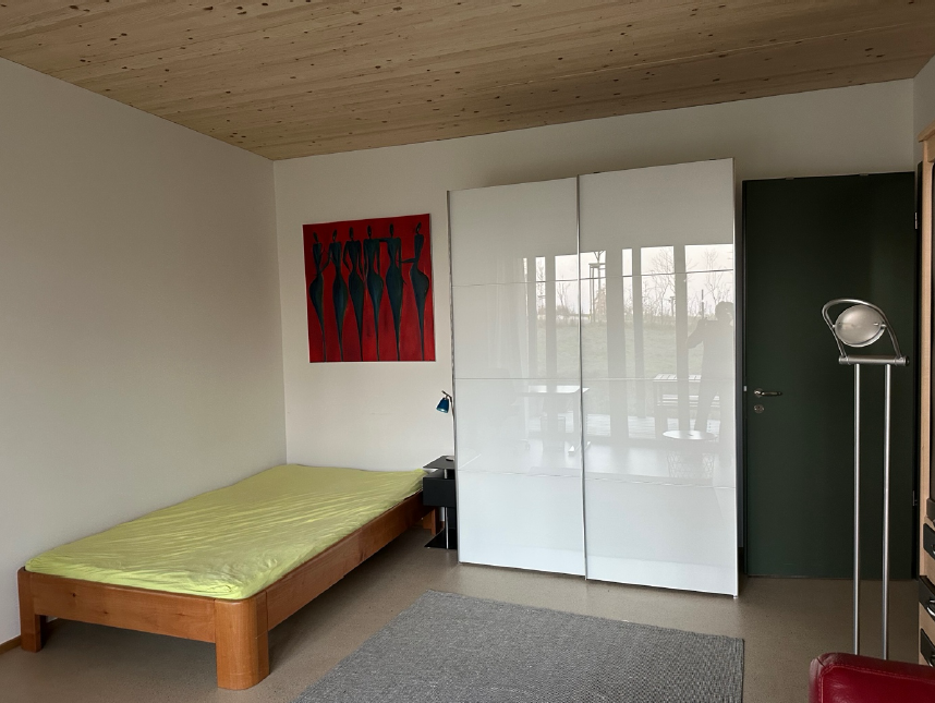 Man sieht ein Einzelbett, einen Kleiderschrank, eine Lampe, sowie die Zimmertüre von innen. Am Boden liegt ein Teppich und an der Wand hängt ein Bild.