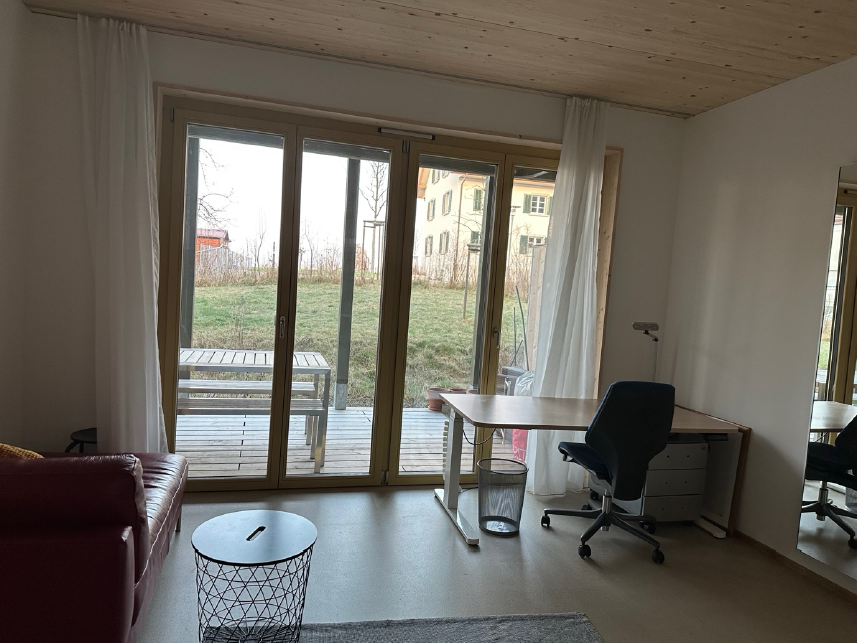 Es ist ein Schreibtisch mit Stuhl, sowie ein Sofa und an der Wand ein Spiegel zu sehen. In der Mitte ist die Türe zum Sitzplatz, an welchen eine Wiese angrenzt.